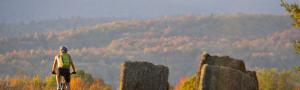 Mountain biker riding through rolling pastural hills with large hay bales and horizon of rolling forested hills of vibrant fall foliage in the Mad River Valley Vermont